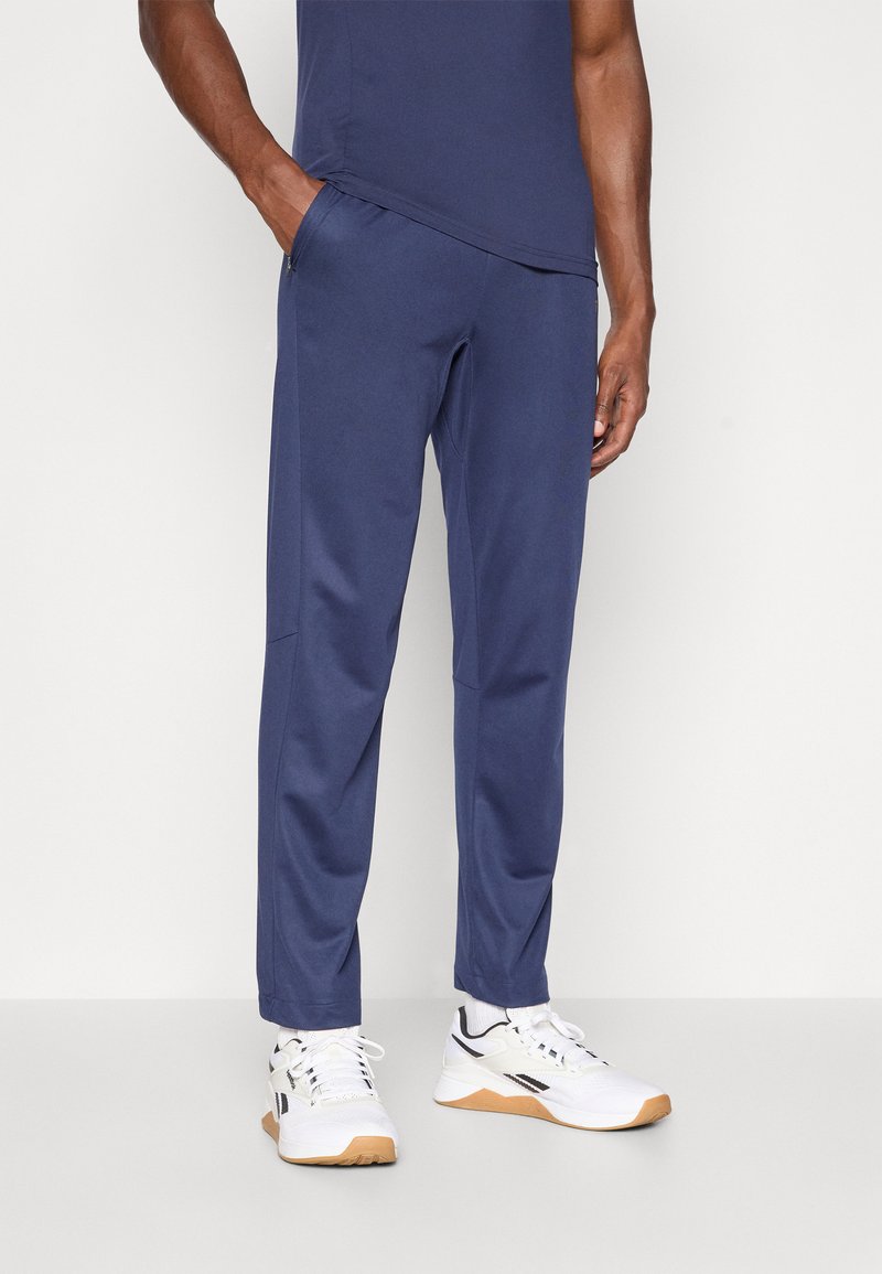 Σκούρες μπλε αθλητικές παντελόνες από μαλακό ύφασμα με πλαινά μπατζάκια, ελαφριά σχεδίαση και στενές γραμμές, συνδυασμένες με λευκά αθλητικά παπούτσια.