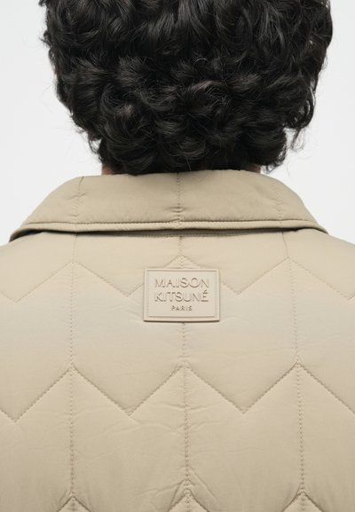Μπεζ καπιτονέ μπουφάν με κωνικό γιακά και δομημένο γεωμετρικό σχέδιο. Μια υφασμάτινη ταυτότητα με λογότυπο είναι ραμμένη στο πίσω μέρος κοντά στην λαιμόκοψη.