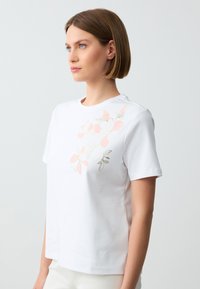 Witte katoenen t-shirt met een bloemenontwerp in zachtroze en goud. Het heeft een ronde hals en korte mouwen, met een losse pasvorm.