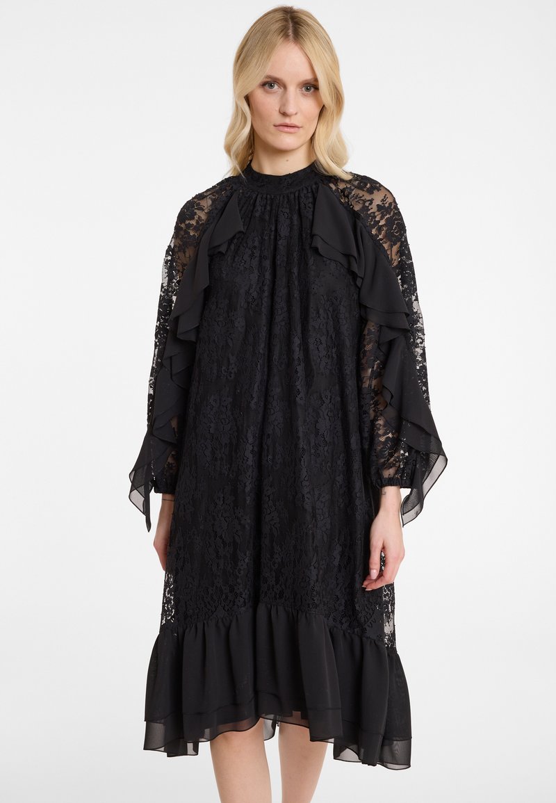 Schwarzes Spitzenkleid mit langen, rüschigen, transparenten Ärmeln, hohem Kragen und ausgestellt geschnittenem Saum mit rüschenförmigem Akzent. Weiche Textur über das gesamte Kleid.