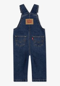 Salopette en denim bleu foncé avec bretelles réglables. Présente une étiquette au dos et deux poches avant avec des coutures contrastantes.