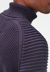 Stickad lila tröja med hög ribbad krage, texturerade mönster på ärmarna och slät finish vid axelsömmen.