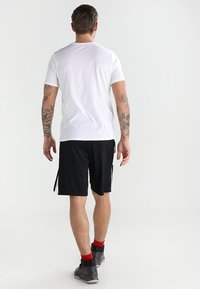 Man med arm-tatueringar som bär en vit t-shirt, svarta shorts, röda strumpor och grå basket skor, står vänd bortåt mot vit bakgrund.