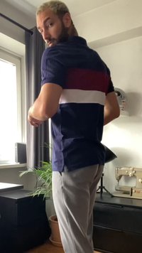Polo shirt blu navy con strisce orizzontali rosse e bianche, abbinato a pantaloni grigi chiari; ambiente interno con piante e mobili visibili.