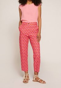 Kvinna som bär en rosa kortärmad topp, högmidjade byxor med geometriskt mönster i rosa och orange, samt beige ankelbandssandaletter.