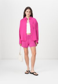 Camicia oversize rosa brillante con bottoni sopra un top bianco corto, abbinata a pantaloni corti rosa testurizzati e infradito nere. Si tiene una piccola borsa bianca.