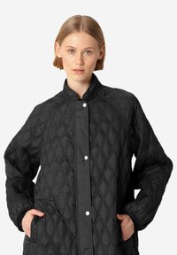 Chaqueta negra acolchada con un cuello alto, que presenta cierres de botones a presión y patrones de costura en forma de diamante. Incluye bolsillos laterales.