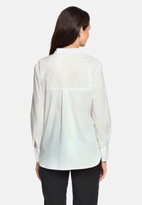 White long-sleeve blouse with a back yoke detail and slight pleat. Features buttoned cuffs and a straight hem. Smooth fabric texture.