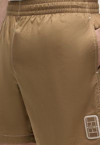 Short marron avec taille élastique, coutures blanches et un petit patch brodé près de l'ourlet représentant un court de tennis et un logo de crocodile.