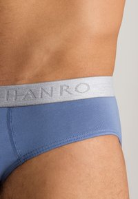 Slips di cotone blu con un elastico argentato in vita, caratterizzati da un logo ricamato. Texture liscia con cuciture in rilievo.
