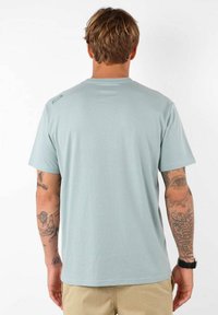 T-shirt en coton bleu clair à manches courtes, avec un col rond et un branding minimal sur l'épaule. Texture douce et coupe décontractée.