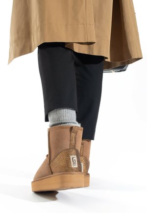 Botines de ante marrón con acentos de purpurina y una etiqueta; combinados con pantalones negros y calcetines grises, mostrando materiales texturizados y un diseño casual.