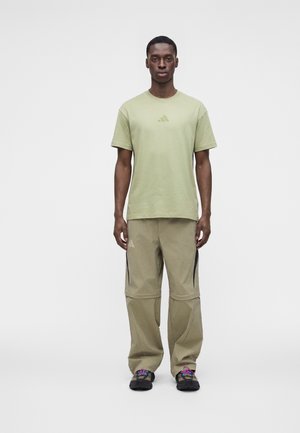Homme debout, face à l'avant, portant un t-shirt Adidas vert clair, un pantalon beige avec détail de fermeture éclair, et des chaussures noires avec des lacets colorés.