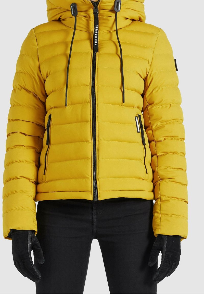 Veste matelassée jaune vif avec fermeture éclair à l'avant, poches zippées, cordons de capuche, portée avec des gants noirs et un pantalon noir.