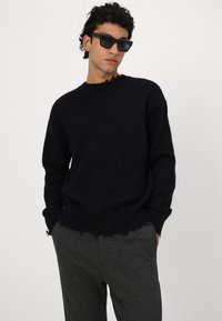 Svart strikkegenser med en avslappet passform, slitt kant og lange ermer, kombinert med grå bukser. Stoffet ser mykt og teksturert ut.