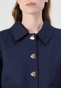 Marineblauwe textuurjas met een spreidkraag, hartvormige gouden knopen en een hartpatroon dat in de stof is gestikt.