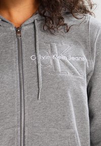 Šedá mikina na zip vyrobená z měkké látky s přední kapsou. Obsahuje vyšívané logo "Calvin Klein Jeans" a kapuci na šňůrku.