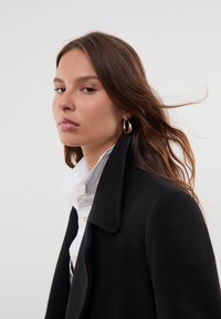 Manteau en laine noir avec un col cranté, associé à une chemise blanche. Présente une texture douce et une coupe décontractée, mise en valeur par des boucles d'oreilles créoles.
