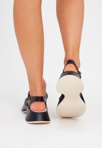 Černé kožené sandály s nastavitelným nártem, nízkým klínovým podpatkem a bílou gumovou podrážkou. Hladký povrch a minimalistický design.