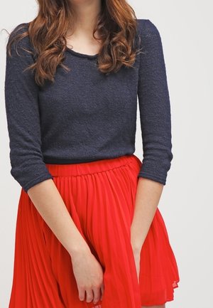 Marineblauwe longsleeve top met een gestructureerde afwerking, gecombineerd met een geplooide rode rok. De rok heeft een elastische tailleband voor extra comfort.