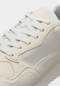 Κοντινό πλάνο ενός εκρού αθλητικού παπουτσιού με υφασμάτινες επιφάνειες με υφή, ραμμένη λεπτομέρεια και λευκά επίπεδα κορδόνια δεμένα σε ζιγκ-ζαγκ.