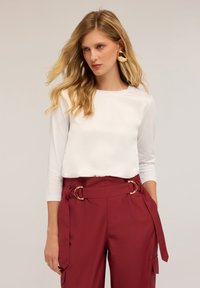 Valkoinen pitkähihaine paita yhdistetty korkeavyötäröisiin burgundinvärisiin housuihin, joissa on vyö kultaisilla yksityiskohdilla ja sivutaskut.