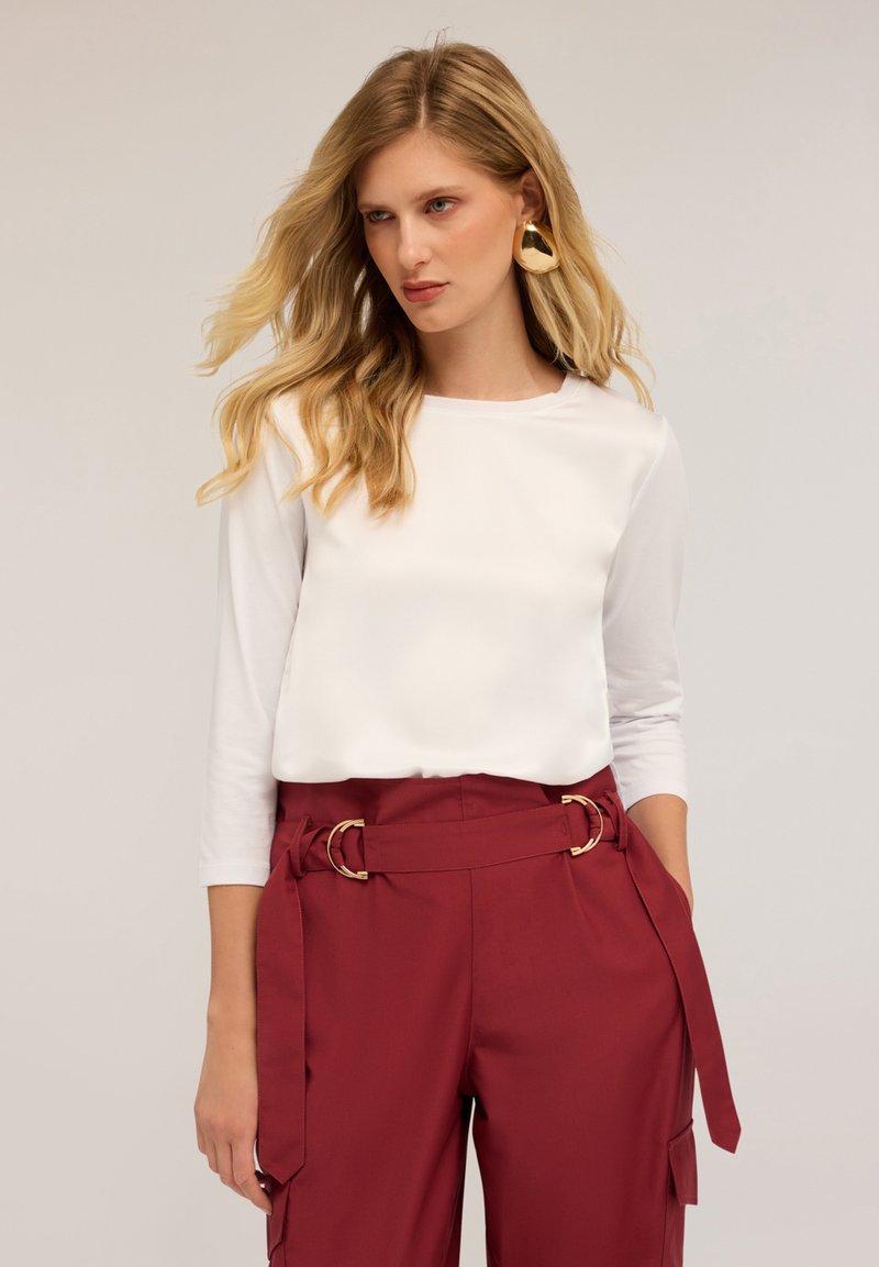 Valkoinen pitkähihaine paita yhdistetty korkeavyötäröisiin burgundinvärisiin housuihin, joissa on vyö kultaisilla yksityiskohdilla ja sivutaskut.