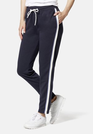 Frau in navy Jogginghosen mit weißen und blauen Seitenstreifen und weißen Sneakers, die mit einer Hand in der Tasche steht.