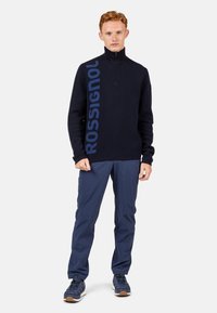 Marinefarbener Fleece mit Reißverschluss und prominenter blauer "ROSSIGNOL"-Logo, kombiniert mit marinefarbenen Hosen und dunklen Sneakers. Schlichtes, funktionales Design.