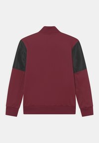 Burgundská bunda s černými saténovými rukávy, zipovým límcem, žebrovanými manžetami a lemem. Hladká textura, atletický design, bez viditelných log nebo vzorů.