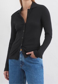 Kvinna iklädd en svart ribbad cardigan med knappar och krage samt blå högmidjade jeans, med ena handen i fickan mot en vit vägg.