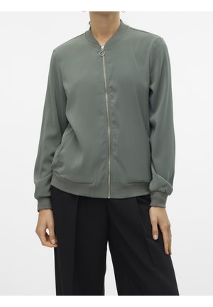 Femme portant une veste bomber zippée vert mat avec col et poignets côtelés, associée à un pantalon noir ample, debout sur fond blanc.