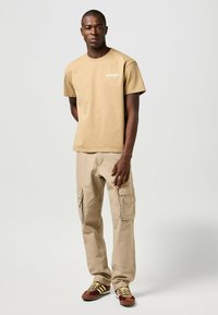 Camiseta de algodón marrón con el logo "Wrangler", combinada con pantalones cargo beige que tienen bolsillos laterales y zapatillas deportivas marrones y amarillas.