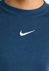 Marineblå hettegenser laget av mykt stoff, med en hvit brodert Nike-logo på brystet og en ribbestrikket halslinning.
