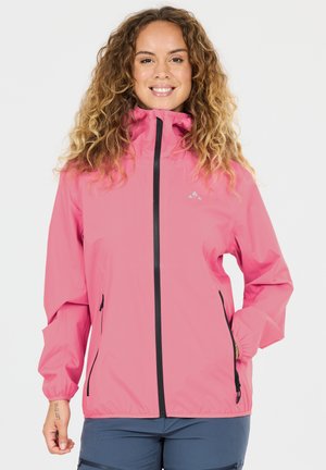 Frau mit lockigem Haar, die eine rosa Reißverschlussjacke und blaue Hose trägt, lächelt mit der rechten Hand in der Jackentasche.