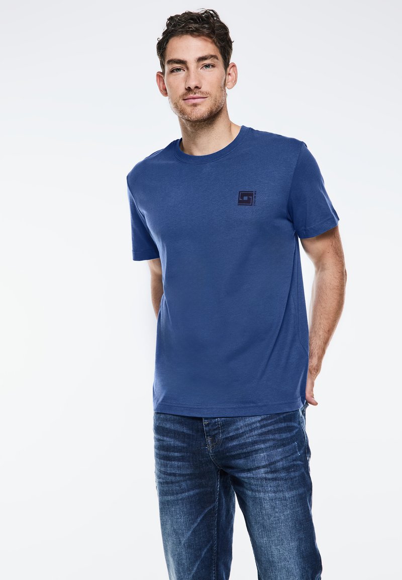 T-shirt di cotone blu con collo rotondo e maniche corte, caratterizzato da un piccolo dettaglio grafico nero sul petto. Indossato con jeans in denim.