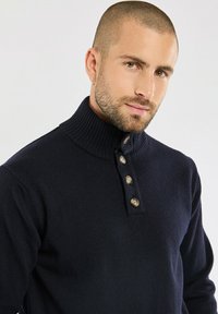 Pull en marine avec col montant à côtes et patte de boutonnage à quatre boutons, fabriqué à partir d'un matériau doux semblable à de la laine, avec un tricot texturé et un design ajusté.