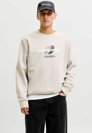 Młody mężczyzna ubrany w beżową bluzę Jack & Jones, czapkę Jack & Jones oraz czarne dżinsy, stojący z jedną ręką w kieszeni.