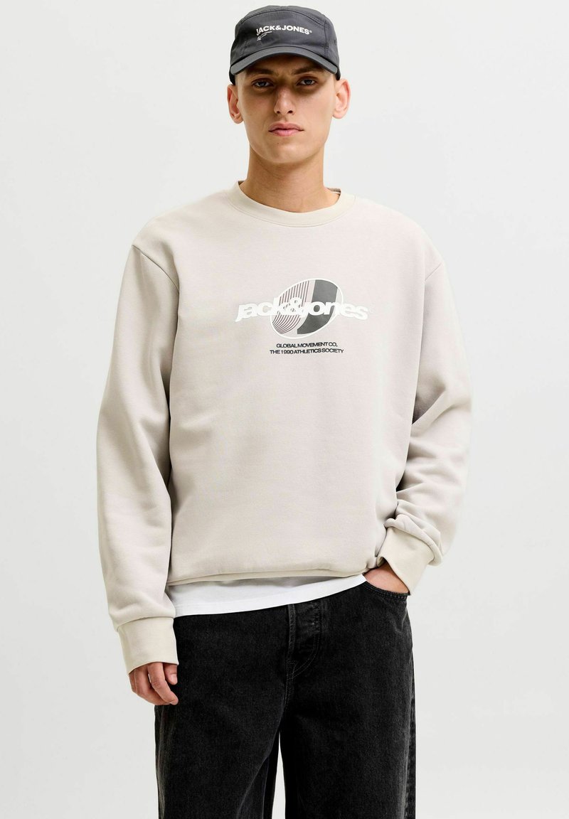 Jeune homme portant un sweat-shirt beige Jack & Jones, une casquette noire Jack & Jones et un jean noir, debout avec une main dans la poche.
