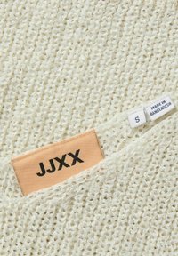 Top de punto texturizado en color blanco sucio, con una etiqueta tejida que muestra "JJXX" y una etiqueta que indica "Hecho en Bangladesh" y tamaño "S".
