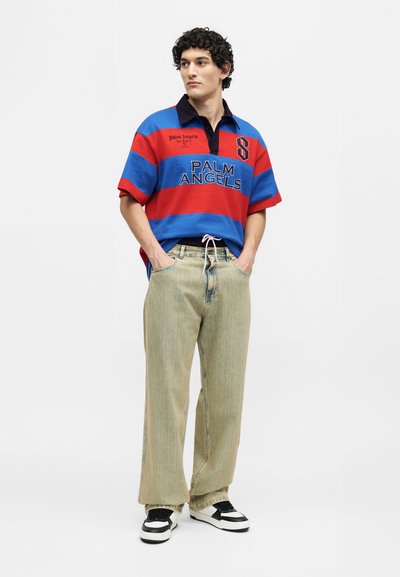 Νέος άνδρας με σγουρά μαλλιά που φοράει μια κόκκινη και μπλε ριγέ ρακέτα Palm Angels, άνετο μπεζ παντελόνι και ασπρόμαυρα αθλητικά παπούτσια, στέκεται με τα χέρια στις τσέπες.