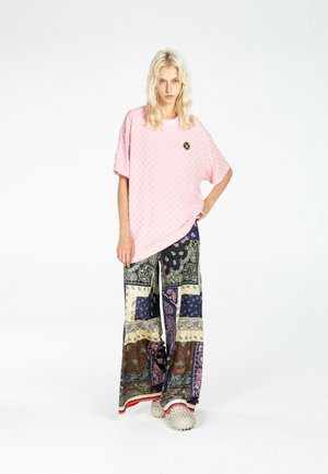 Mujer joven con pelo largo y rubio que lleva una camisa rosa de cuadros extragrande y pantalones anchos de patchwork con detalles estampados.