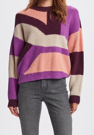 Femme portant un pull tricoté multicolore avec des motifs abstraits rose, violet, bordeaux et beige, et un jean gris taille haute.