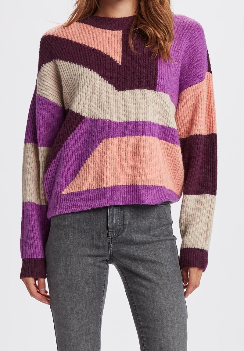 Femme portant un pull tricoté multicolore avec des motifs abstraits rose, violet, bordeaux et beige, et un jean gris taille haute.
