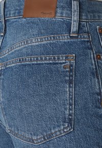 Blue denim jeans featuring a brown leather waistband label, exposed stitching, and back pocket with a small logo detail.