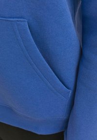 Blaue Sweatshirt mit strukturiertem Stoff, ausgestattet mit einer Fronttasche und gerippten Bündchen. Nahaufnahme, die die Stoffstruktur und die Farbe betont.