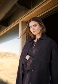 Mørk ruskindsjakke med krave og sølvknapper, lagdelt over en gennemsigtig, lysebrune bluse. Baggrunden har en træstruktur og naturlandskab.