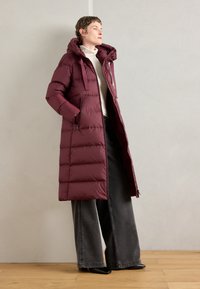 Burgundia puffer jope, millel on kapuuts, millel on tikitud disain, lukuga taskud ja pikad varrukad, koos laia lõikega hallide teksadega.