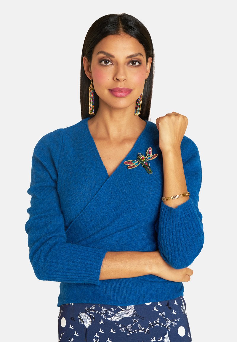 Blaues Wickeloberteil aus weichem, strukturiertem Strick mit gerippten Ärmeln. Mit einem bunten Libellenbrosche. Kombiniert mit einem gemusterten marineblauen Rock.