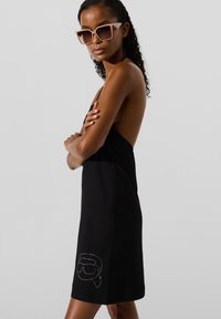 Göndör hajú nő túlméretezett négyzet alakú napszemüvegben, fekete hát nélküli ruhában, amelynek szoknyáján szegecselt arc dizájn látható.